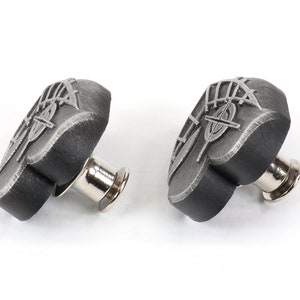 K&ouml;nnte beinhalten: Zwei schwarze herzf&ouml;rmige Metallkn&ouml;pfe mit silbernem Design. Die Kn&ouml;pfe haben einen Stift auf der R&uuml;ckseite zum Anbringen an Stoff.