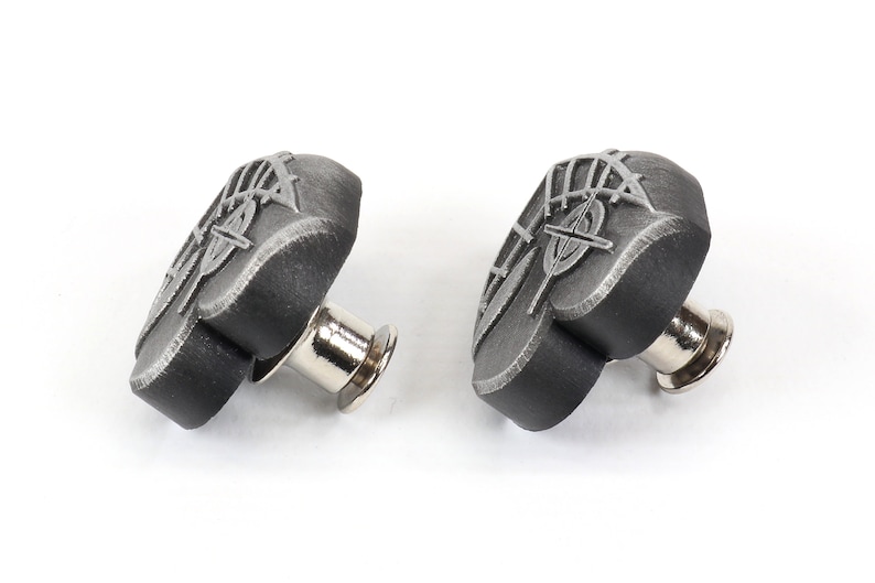 K&ouml;nnte beinhalten: Zwei schwarze herzf&ouml;rmige Metallkn&ouml;pfe mit silbernem Design. Die Kn&ouml;pfe haben einen Stift auf der R&uuml;ckseite zum Anbringen an Stoff.