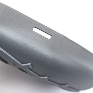 May include: A gray, curved plastic object with a textured surface and a rectangular cutout. The object has a series of decorative cutouts along its edge. The item is likely a component or part of a larger product.