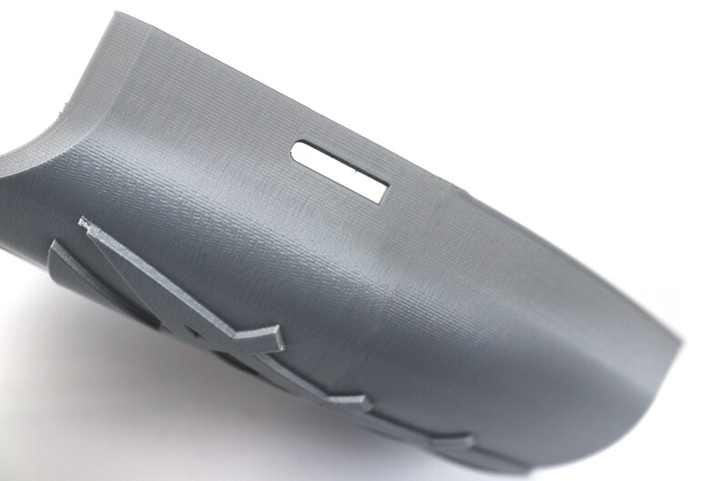 May include: A gray, curved plastic object with a textured surface and a rectangular cutout. The object has a series of decorative cutouts along its edge. The item is likely a component or part of a larger product.