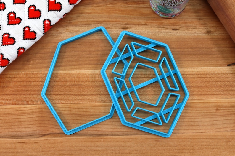 Op de afbeelding: Twee blauwe plastic koekjesvormen in de vorm van zeshoeken. De grotere vorm heeft een geometrisch ontwerp in de zeshoek.