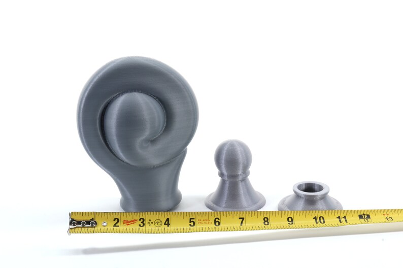 K&ouml;nnte beinhalten: Drei graue 3D-gedruckte Objekte unterschiedlicher Formen und Gr&ouml;&szlig;en werden vor einem wei&szlig;en Hintergrund gezeigt. Ein gelbes Ma&szlig;band liegt neben den Objekten. Das gr&ouml;&szlig;te Objekt hat ein spiralf&ouml;rmiges Design, die anderen beiden sind kleiner und haben eine abgerundete Form.