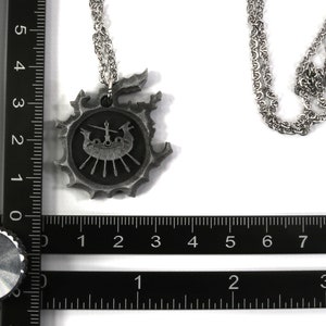 May include: A silver necklace with a pendant featuring a Viking ship with oars and a figurehead. The pendant is surrounded by a decorative border. The necklace is on a black ruler for size reference.