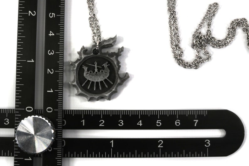 May include: A silver necklace with a pendant featuring a Viking ship with oars and a figurehead. The pendant is surrounded by a decorative border. The necklace is on a black ruler for size reference.