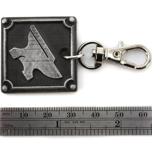 May include: A silver metal keychain with a square charm featuring a stylized anvil design. The charm is attached to a silver metal lobster clasp.
