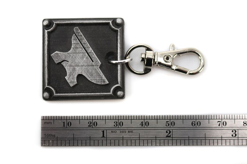 May include: A silver metal keychain with a square charm featuring a stylized anvil design. The charm is attached to a silver metal lobster clasp.