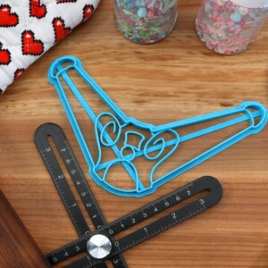 May include: A light blue cookie cutter in the shape of a bell, along with a black angle ruler, a wooden rolling pin, and a blue spatula. Sprinkles and a heart-patterned oven mitt are also visible on a wooden surface.