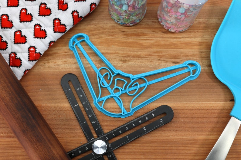 May include: A light blue cookie cutter in the shape of a bell, along with a black angle ruler, a wooden rolling pin, and a blue spatula. Sprinkles and a heart-patterned oven mitt are also visible on a wooden surface.