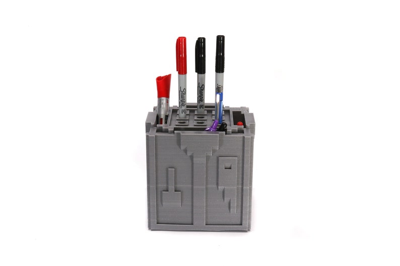 May include: A gray 3D printed pen holder with four compartments. The holder has a red, black, and blue marker in it. The holder is on a white background.