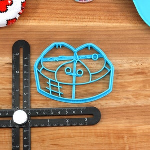 May include: A light blue plastic cookie cutter in the shape of a face, with two eye-like circles and a mouth-like shape. A black ruler with white markings is next to the cutter. Other items are visible in the background.