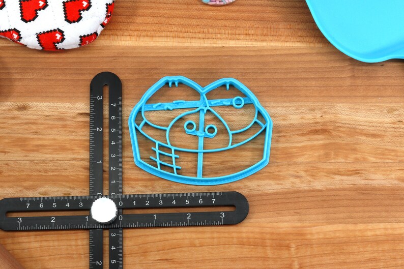 May include: A light blue plastic cookie cutter in the shape of a face, with two eye-like circles and a mouth-like shape. A black ruler with white markings is next to the cutter. Other items are visible in the background.