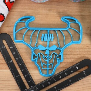 May include: A blue plastic cookie cutter shaped like a skull with horns. The cutter is on a wooden surface with a ruler.
