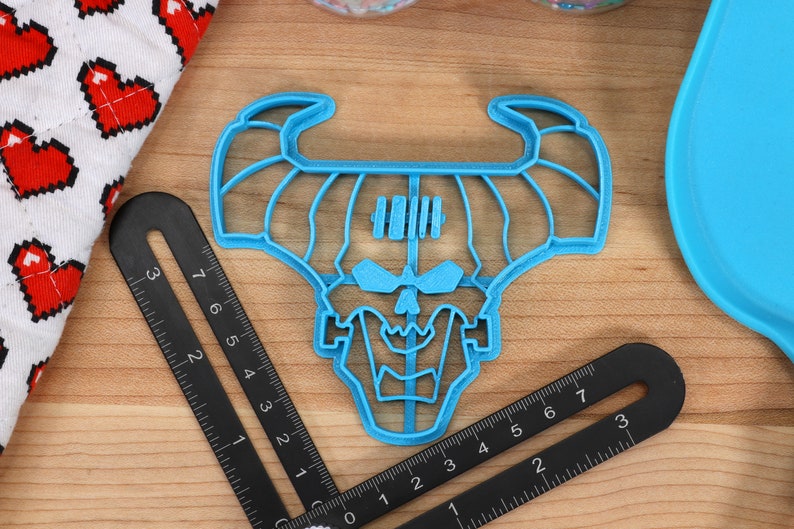 May include: A blue plastic cookie cutter shaped like a skull with horns. The cutter is on a wooden surface with a ruler.