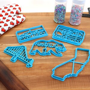 May include: A set of five blue plastic cookie cutters shaped like the Golden Gate Bridge, a California bear, the state of California, a palm tree, and a mountain range. The cutters are on a wooden surface with a rolling pin, a spatula, and two jars of sprinkles.