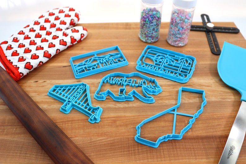May include: A set of five blue plastic cookie cutters shaped like the Golden Gate Bridge, a California bear, the state of California, a palm tree, and a mountain range. The cutters are on a wooden surface with a rolling pin, a spatula, and two jars of sprinkles.