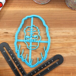 May include: A blue plastic cookie cutter shaped like a gorilla's face. The cookie cutter is on a wooden surface.