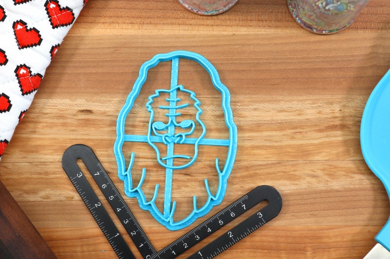 May include: A blue plastic cookie cutter shaped like a gorilla's face. The cookie cutter is on a wooden surface.