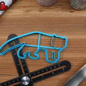 May include: A blue plastic cookie cutter shaped like a bear. The cutter is lying on a wooden surface with a ruler and a blue spatula.