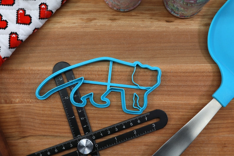 May include: A blue plastic cookie cutter shaped like a bear. The cutter is lying on a wooden surface with a ruler and a blue spatula.