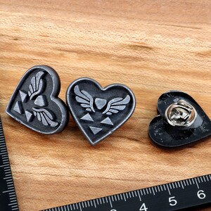 Puede incluir: Tres pines de coraz&oacute;n plateados con un s&iacute;mbolo alado y un dise&ntilde;o triangular. Los pines est&aacute;n sobre una superficie de madera.