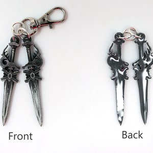 May include: A silver-colored keychain featuring two decorative dagger charms. The front side of the daggers has intricate detailing, while the back side is smooth. The keychain includes a lobster clasp for easy attachment.