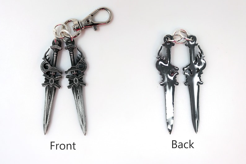May include: A silver-colored keychain featuring two decorative dagger charms. The front side of the daggers has intricate detailing, while the back side is smooth. The keychain includes a lobster clasp for easy attachment.