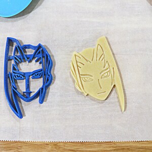 May include: Two cookie cutters, one blue and one beige, shaped like human faces with long hair. The blue cutter has a more serious expression, while the beige cutter has a more neutral expression.