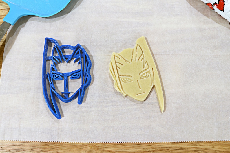 May include: Two cookie cutters, one blue and one beige, shaped like human faces with long hair. The blue cutter has a more serious expression, while the beige cutter has a more neutral expression.