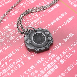 Puede incluir: Un collar de plata con un colgante con forma de dispositivo digital. El colgante tiene una pantalla negra y un marco plateado con tres botones. El collar est&aacute; sobre una superficie rosa con texto japon&eacute;s.