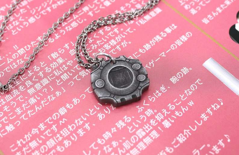 Puede incluir: Un collar de plata con un colgante con forma de dispositivo digital. El colgante tiene una pantalla negra y un marco plateado con tres botones. El collar est&aacute; sobre una superficie rosa con texto japon&eacute;s.