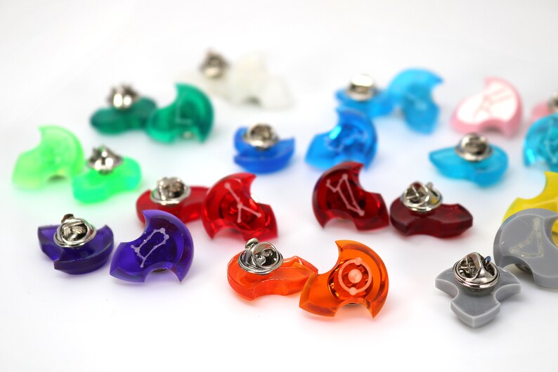 May include: A collection of colorful, crescent-shaped pins with zodiac symbols. The pins are made of clear plastic and have a silver pin back.
