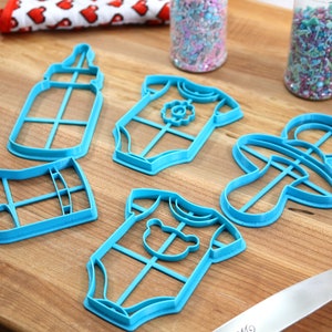 May include: A set of six blue plastic cookie cutters shaped like a baby bottle, a baby onesie, a pacifier, and a sock. The cutters are on a wooden surface.