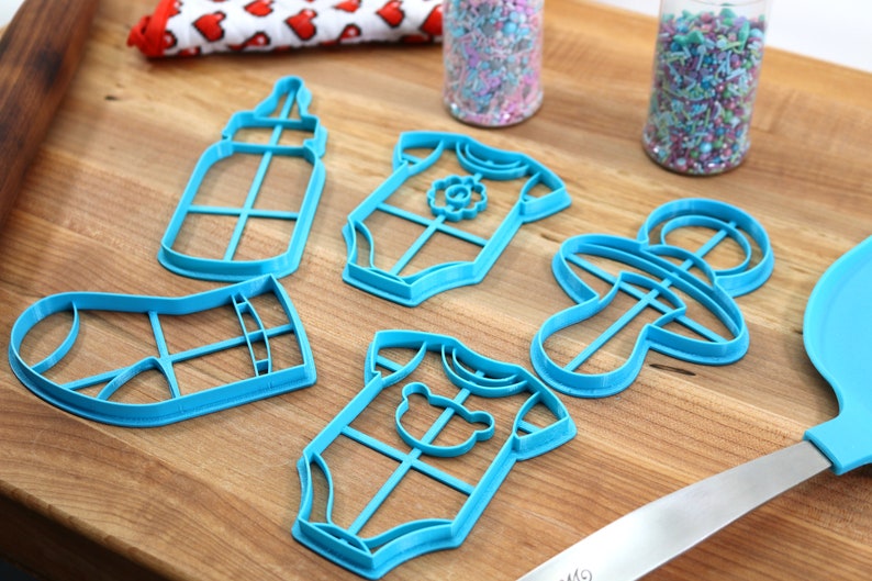 May include: A set of six blue plastic cookie cutters shaped like a baby bottle, a baby onesie, a pacifier, and a sock. The cutters are on a wooden surface.