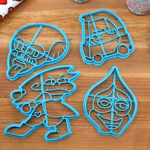 Puede incluir: Conjunto de cuatro cortadores de galletas de pl&aacute;stico azul con forma de caras de dibujos animados y un coche. Los cortadores est&aacute;n sobre una superficie de madera.