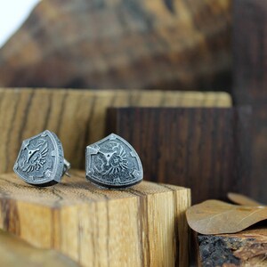 May include: A pair of silver shield-shaped earrings with a detailed design. The earrings are displayed on a wooden surface.