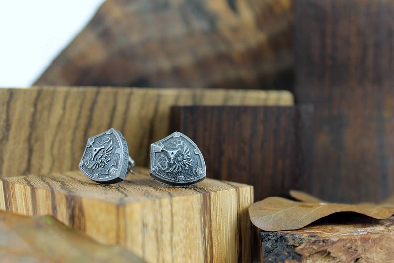 May include: A pair of silver shield-shaped earrings with a detailed design. The earrings are displayed on a wooden surface.