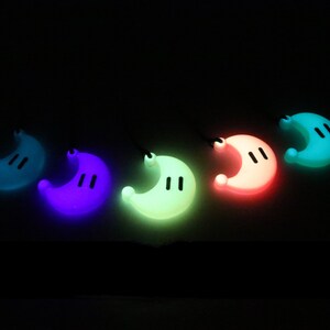 May include: Five glow-in-the-dark crescent moon charms in different colors: teal, purple, green, pink, and blue. Each charm has two small black dots for eyes.
