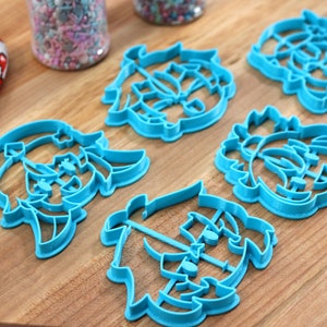 May include: Five blue plastic cookie cutters shaped like cartoon faces. The cutters are on a wooden surface.