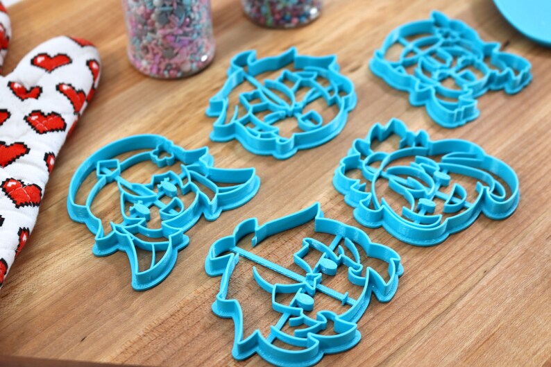 May include: Five blue plastic cookie cutters shaped like cartoon faces. The cutters are on a wooden surface.
