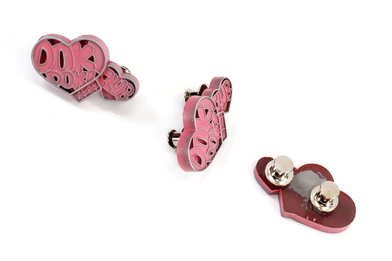 May include: Three pink heart-shaped pins with the text "DOK DOK CLUB" on them. The pins have silver metal backs with a pin and a safety clasp.