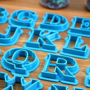 May include: A set of blue plastic alphabet cookie cutters. The cutters are arranged on a wooden surface.