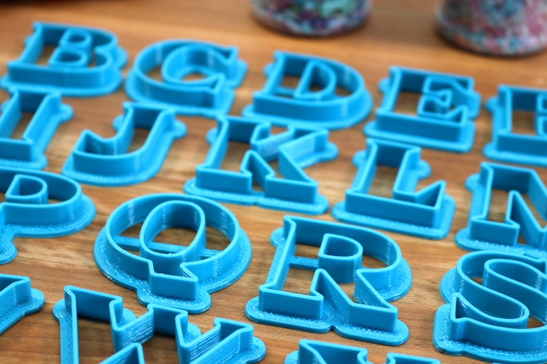 May include: A set of blue plastic alphabet cookie cutters. The cutters are arranged on a wooden surface.