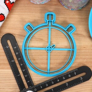 May include: A blue plastic cookie cutter shaped like a stopwatch. The cutter is on a wooden surface with a ruler and other baking supplies.