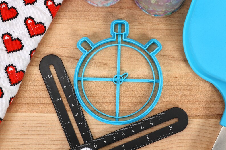 May include: A blue plastic cookie cutter shaped like a stopwatch. The cutter is on a wooden surface with a ruler and other baking supplies.