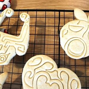 May include: Five cookies on a wire rack. The cookies are decorated with different designs, including a heart, a pineapple, and a geometric shape. The cookies are light brown in color.