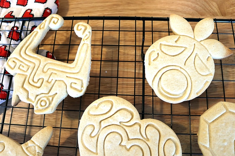May include: Five cookies on a wire rack. The cookies are decorated with different designs, including a heart, a pineapple, and a geometric shape. The cookies are light brown in color.