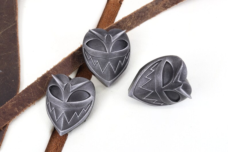 May include: Three silver heart-shaped pins with a menacing cartoon face design. The pins have a black and silver color scheme.