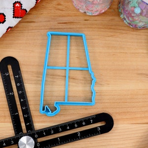 May include: A blue plastic cookie cutter shaped like the state of Alabama. The cutter is on a wooden surface with a ruler and other baking supplies.
