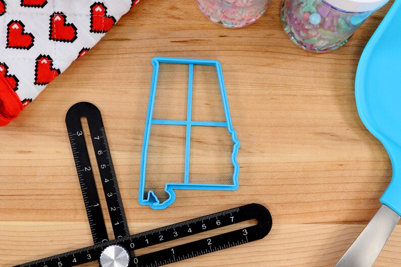 May include: A blue plastic cookie cutter shaped like the state of Alabama. The cutter is on a wooden surface with a ruler and other baking supplies.