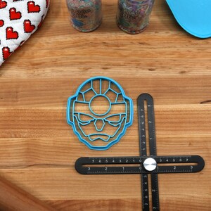May include: A blue plastic cookie cutter shaped like a cartoon face with a large circle in the center of the face. The cookie cutter is on a wooden surface with a black ruler and a rolling pin.